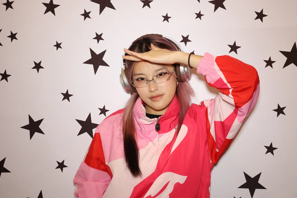 Emily with pink hair, silver headphones and a pink jacket in front of a starry backdrop.